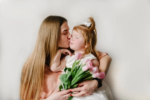 The mother kissing little girl on light background indoor. Portrait of mom with flowers and daughter sitting in home. The concept of happy family holiday. woman\'s day. Mother\'s Day.