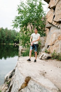 Bir adam büyük bir taşın arkasındaki büyük kayaların arkasına gider. Tam boy, aşağıdan görüş. Kameraya bakıyorum. Kapatın. Metin ve tasarım için yer. Adam göl kenarındaki uçurumda poz veriyor..