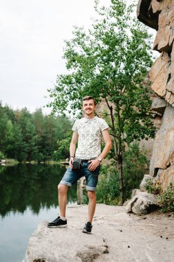 Bir adam büyük bir taşın üzerindeki büyük kayaların arkasında duruyor. Tam boy, aşağıdan görüş. Kameraya bakıyorum. Kapatın. Metin ve tasarım için yer. Adam göl kenarındaki uçurumda poz veriyor..