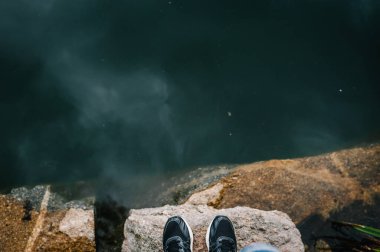 Erkeklerin bacakları gölün önündeki bir taşın üzerinde duruyor. Suya taşları taşırdı. Nehre atlamadan önce. Spor ayakkabılar. Kapatın. Üst Manzara. taş.