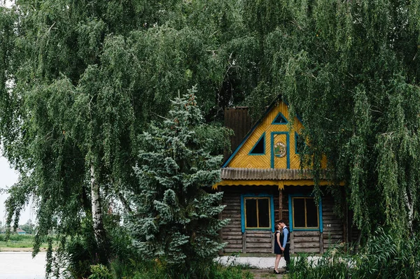 一对年轻夫妇在森林间的一座岛上的一座木制老房子旁边站着拥抱和亲吻 房子在树后 大自然 靠近点 — 图库照片