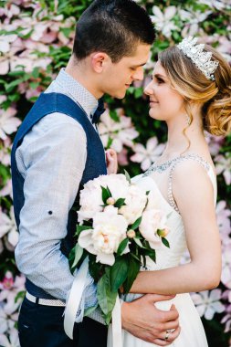 Taçlı ve damatlı şakayık desenli şık bir gelin. Koca, kadının belinin arkasına sarılır. Kapatın. Clematis. Çiçekler nazikçe pembe, mor ve yeşil yapraklardır. Çalılar, ağaçlar.