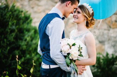 Bir buket şakayık ve damat buketi ile şık, mutlu bir gelin. Ellerinde balonlarla sarılıyorlar. Evin avlusundaki doğanın arka planında. Kapatın. taş duvar.