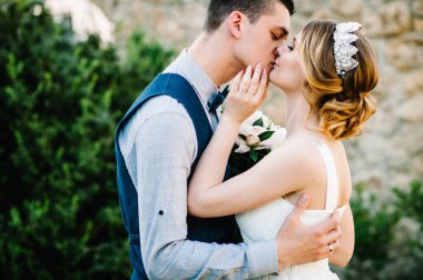 Taçlı ve damatlı şakayık desenli şık bir gelin. Koca belinin arkasından sarılır ve kadını öper. Kapatın. Doğa. Taş duvarın arkasında..