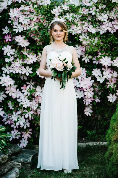 Stylish portrait bride with bouquet of peonies with crown. Close up. Clematis. flowers are pink and purple, green leaves. bushes. Charming, serious, handsome young girl isolated on background nature