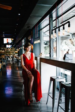 Kafe masasının penceresinde oturan akşam kırmızısı elbiseli güzel, seksi Avrupalı esmer kız. Kameraya ciddi ciddi bakıyorsun. Pozlar. Tüm büyüme.