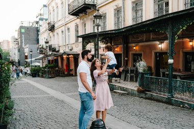 Genç mutlu aile: baba, kız, anne eski şehrin, kasabanın, açık havanın antik mimarisinin yakınındaki bir sokakta yürürken bir bebek fırlatıyor. Aile tatili ve seyahat kavramı.