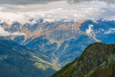 Dağ manzarası. Sıcak sonbahar güneşli bir gün. Dağlarındaki sonbahar ormanları ile kaplı. Bulutlar dağ zirveleri yukarıda. Üstleri karda