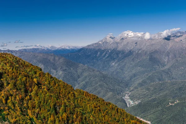 Dağ manzarası. Sıcak sonbahar güneşli bir gün. Dağlarındaki sonbahar ormanları ile kaplı. Bulutlar dağ zirveleri yukarıda. Üstleri karda