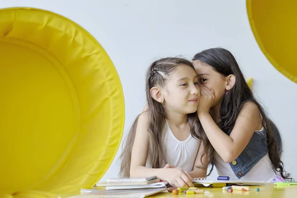 Girl whispering a secret to another girl\'s ear in a learning space.