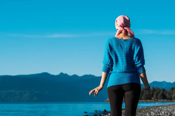 A woman with cancer on her back wearing a pink scarf head and a blue coat is looking at the lake on a beautiful day