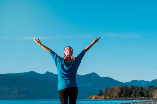 Woman with cancer wearing a pink head scarf and a blue coat with her arms open while looking at the sky and the lake on a beautiful day