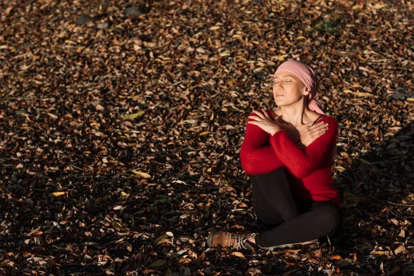 A woman with cancer is sitting hugging herself, smiling and taking the sunshine in the meadow with dry leaves. She wears a pink headscarf and a red coat.