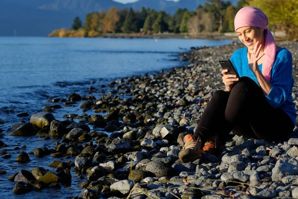 Woman with cancer wearing a pink headscarf is in a video call with her cell phone sitting on the beach during a sunset