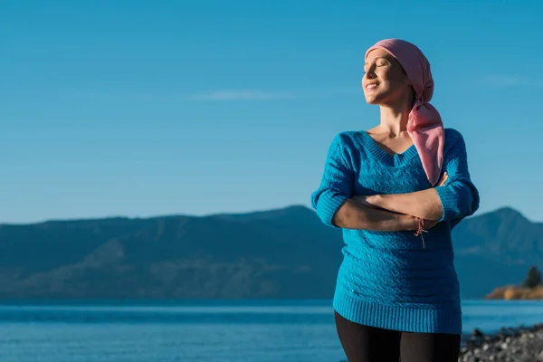 A woman with cancer wears a pink scarf on her head and stands with her arms crossed and smiling into the sun.