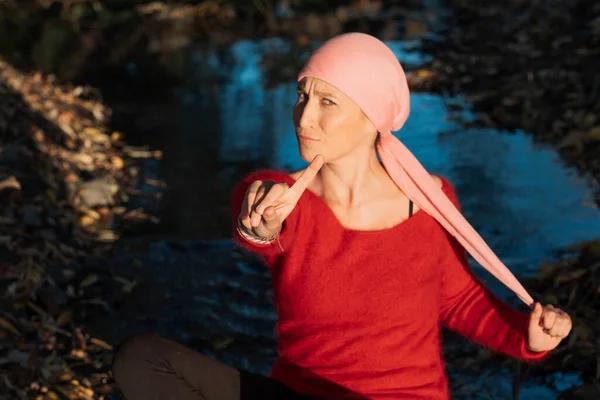 A woman with cancer looks at the camera and gestures her hand saying NO in a park. She wears a pink scarf and a red blouse