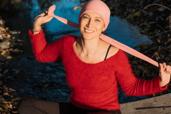 Woman with cancer looks at the camera and smiles as she ties her pink scarf head