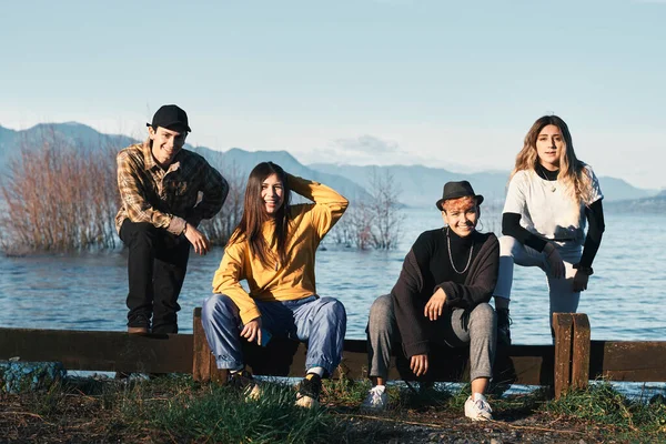 Group of millennial friends near a lake are having fun posing for pictures looking at the camera. Modern youth fashion