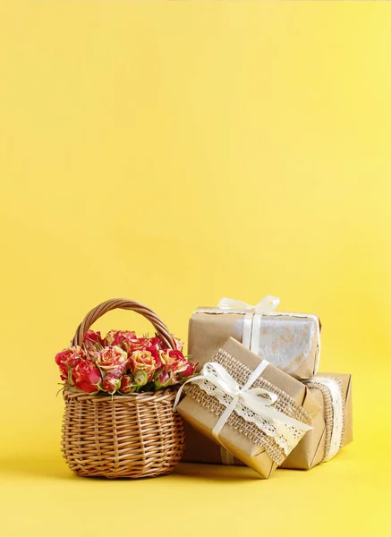 Wicker korg med rosor och vackert inslagna gåvor på gul — Stockfoto