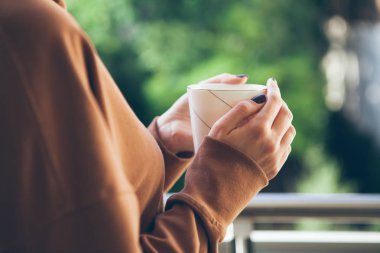 Kadın bir balkonda kahve içiyor. Ellerin beyaz fincan çay ile kapatın. Sabah rutin. Seçici odak. Yeşil arka plan bulanık.