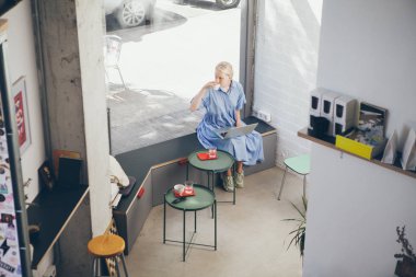 İskandinav tarzı bir kafede kahve içen genç bir kadının en iyi fotoğrafı. Mavi elbiseli kadın dizüstü bilgisayar kullanır ve ofis dışında kahve içer.