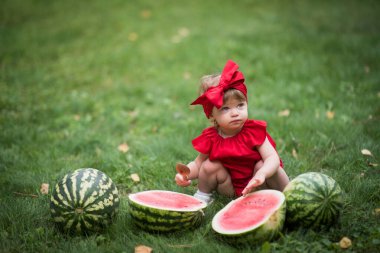 Küçük kız yeşil çimlere oturup kırmızı elbiseyle karpuz yiyor. 