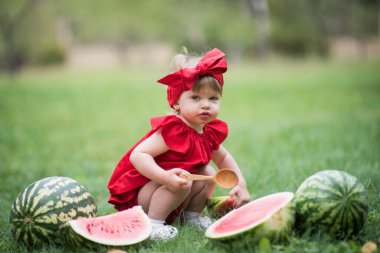 Küçük kız yeşil çimlere oturup kırmızı elbiseyle karpuz yiyor. 