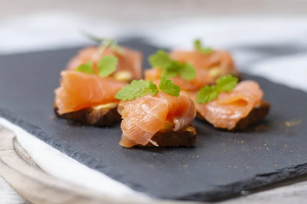 Salmon asap pada roti panggang dengan mentega dengan daun mint di batu hitam dan papan kayu tua - Gambar — Stok Foto