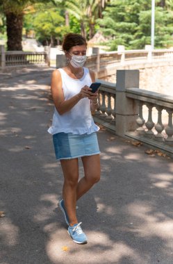 Tıbbi maskeli bir kadın akıllı telefon kullanıyor, parkta yürüyor.