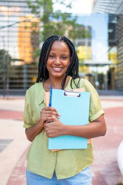 Üniversite kampüsünde elinde dosyalar ve kalemle gülümseyen Afrikalı Amerikalı bir öğrencinin portresi.