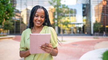 Kentsel finans bölgesinde gülümseyen ve tablet kullanan genç yönetici, arka planda modern binalar