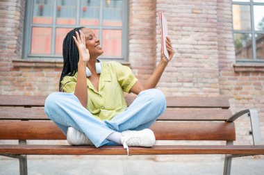 Genç siyahi profesyonel kadın bir bankta oturup ekranda el sallarken tabletle video görüşmesi yapıyor.