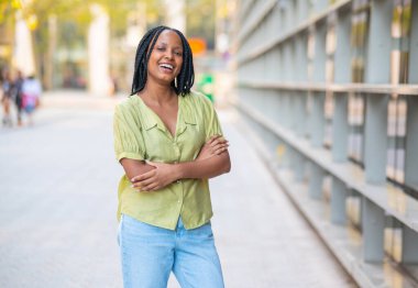 Şehir ortamında kollarını kavuşturmuş gülümseyen genç bir zenci kadının portresi.