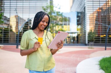 Gülümseyen Afrikalı Amerikalı kadın şehirde online alışveriş için dijital tablet ve kredi kartı kullanıyor.