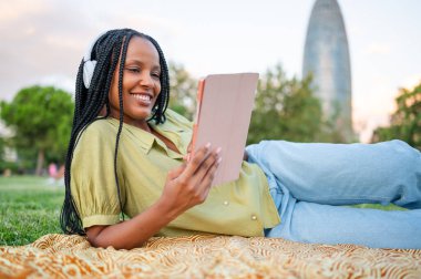 Tablet kullanarak battaniyeye uzanan ve parkta kulaklık takan genç bir kadın.
