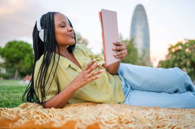 Kulaklık takan ve tablet kullanan genç bir kadın Barselona 'da bir parkta battaniyeye uzanmış video çağrısı yapıyor.