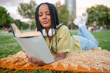 Genç bir kadın parkta dinleniyor kitap okuyor ve kulaklıkla müzik dinliyor.