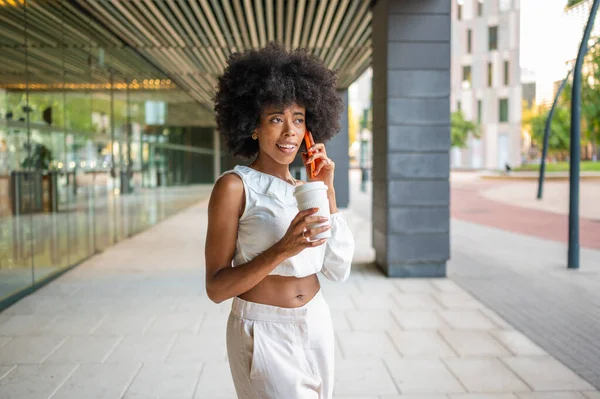 Young black businesswoman walking in the city and talking on the phone while holding a coffee