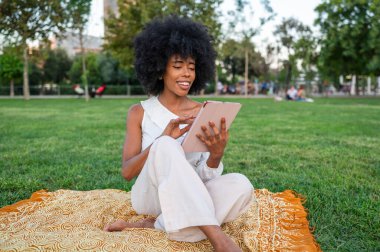 Young woman using digital tablet while sitting on blanket in a park, enjoying leisure time on a sunny day