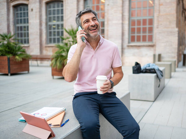 Smiling man talking on phone, holding coffee cup in urban outdoor setting