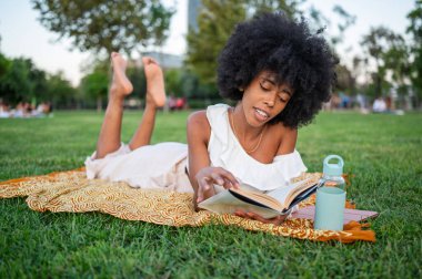 Young woman enjoying reading a book while lying on a blanket in a park, during a sunny day