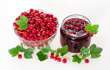 Bir kavanoz ve bir cam fincan beyaz zemin üzerine yeşil yaprakları ile olgun meyveleri kırmızı kuş üzümü reçeli. Üstten görünüm.