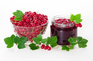 Bir kavanoz ve bir cam fincan beyaz zemin üzerine yeşil yaprakları ile olgun meyveleri kırmızı kuş üzümü reçeli.