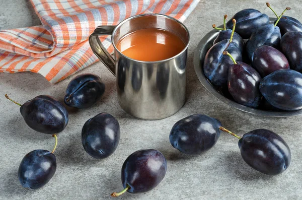 Plum juice in a metal mug and ripe plums in a plate on a gray table.