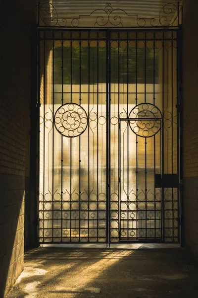 High contrast old metal door gate with many details in sepia tone surrounded by a stone wall. Light falls through a high iron gate