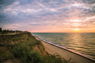 Bulutlu bir sabahta denizde güzel bir gün doğumu. Kızlar bir bankta oturur ve kumlu sahil şeridinde güneşin doğuşunu selamlarlar. Karadeniz. Sanzhiika. Ukrayna