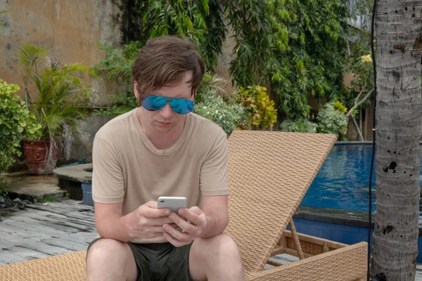 A young man in sunglasses works on the phone against the backdrop of a pool in a tropical country. Freelancer traveler works.