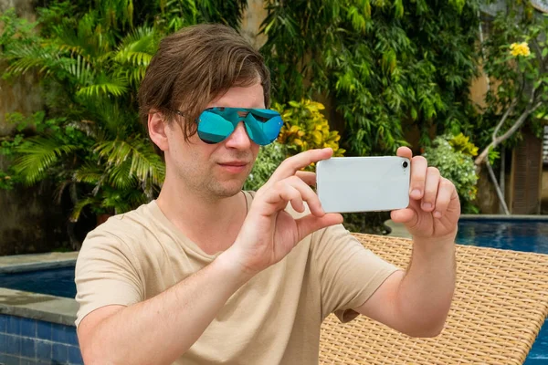 A young man in sunglasses works on the phone against the backdrop of a pool in a tropical country. Freelancer traveler works. Blogger takes content