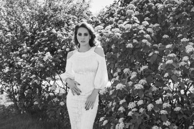 Güzel romantik bir bayanın açık hava portresi. Mükemmel makyaj ve saç stili. Çiçeklerin arkasında poz veren beyaz elbiseli bir kadın..