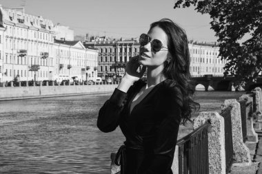 Güzel romantik bir bayanın açık hava portresi. Mükemmel makyaj ve saç stili. Dalgalı saçlı, güneş gözlüklü ve siyah kısa elbiseli bir esmer sahildeki köprüde duruyor..